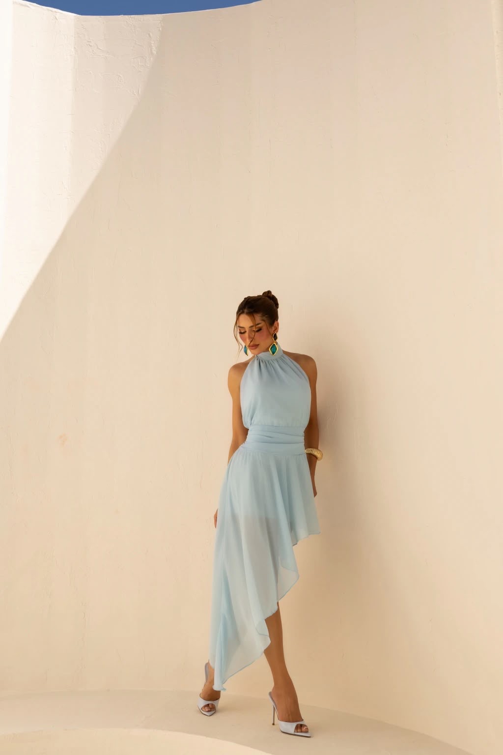 Modelo Alba em vestido azul frente a parede curva em tom areia